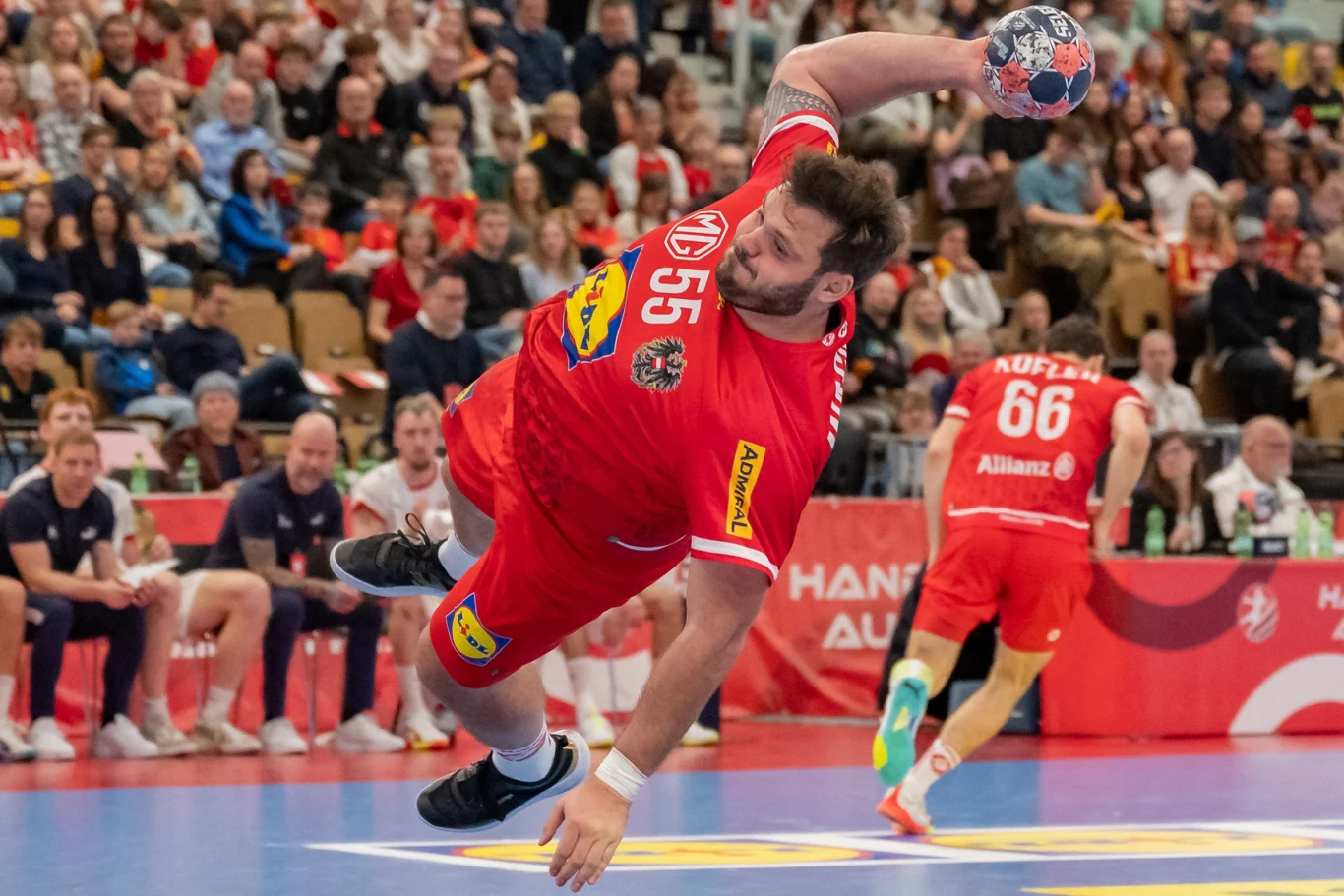 Zawodnik w czerwonym stroju wykonuje dynamiczny rzut piłką ręczną podczas meczu, unosząc się w powietrzu tuż przed oddaniem strzału, z widoczną skupioną widownią w tle.
