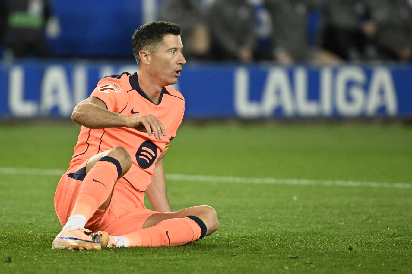 Robert Lewandowski Piłkarz w pomarańczowym stroju Barcelony siedzi na murawie boiska podczas meczu, z wyrazem zaskoczenia lub dezaprobaty na twarzy, w tle fragmenty trybun i napis LaLiga.