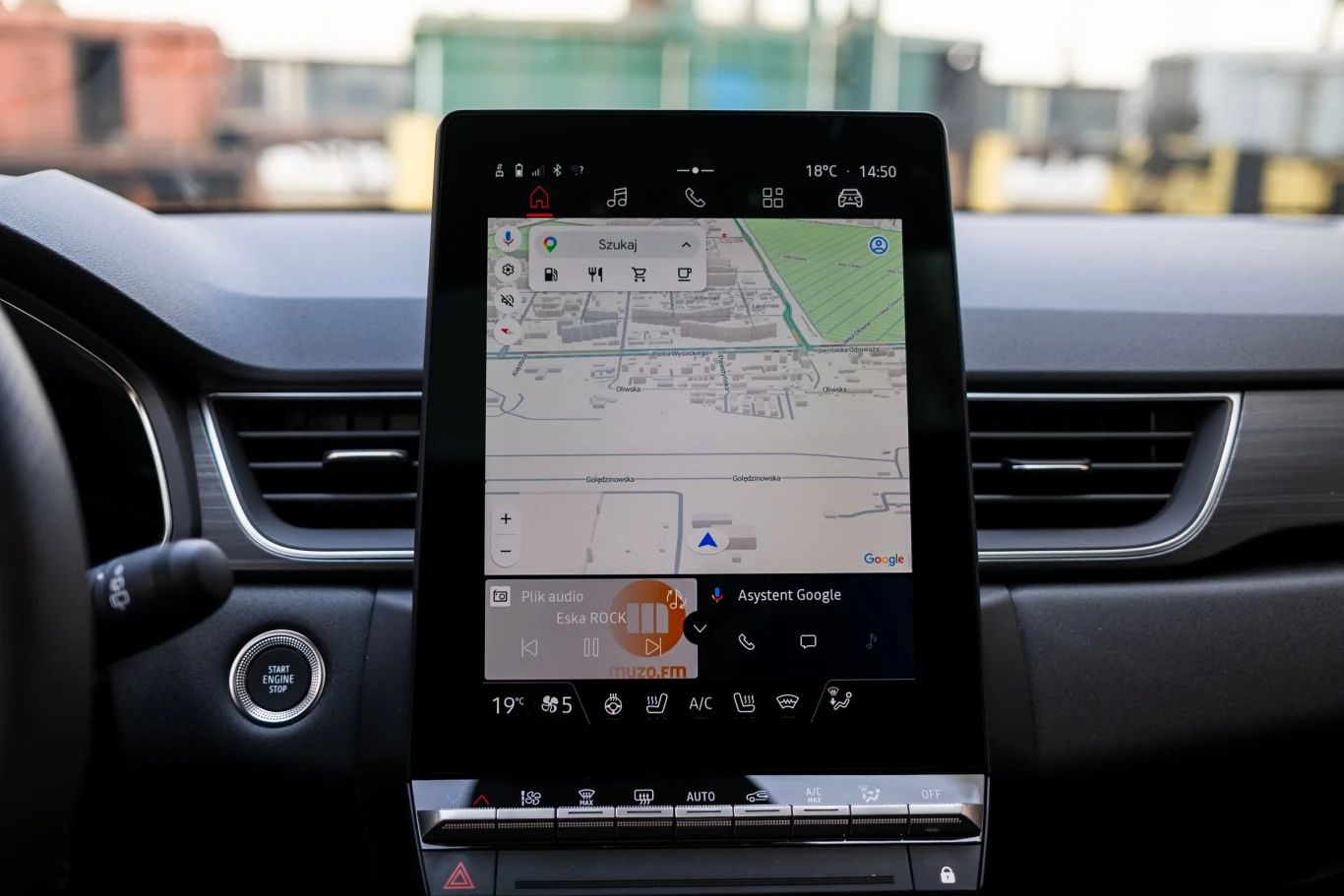 Wbudowany system Google jest dostępny tylko w najwyższej specyfikacji. W niższych nawigacja jest dostępna przez Apple CarPlay i Android Auto. Dotykowy ekran systemu multimedialnego w samochodzie wyświetlający mapę nawigacji, widoczne przyciski kontroli klimatyzacji i panel startowy po lewej stronie.