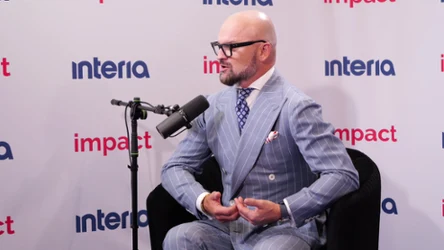 Mężczyzna w eleganckim, niebieskim prążkowanym garniturze rozmawia do mikrofonu studyjnego, siedząc na czarnym fotelu na tle ściany z logo interia i impact.