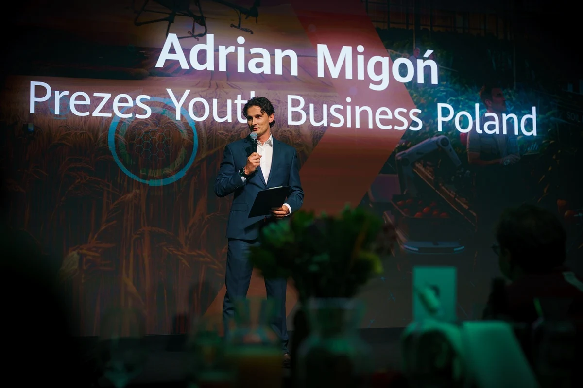 Mężczyzna w garniturze przemawia na scenie z mikrofonem w dłoni, na tle dużego ekranu z napisem „Adrian Migoń Prezes Youth Business Poland”.