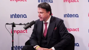 Elegancko ubrany mężczyzna w garniturze i krawacie siedzi na czarnym fotelu i udziela wywiadu do mikrofonu w studiu z logotypami „interia” i „impact” na tle.
