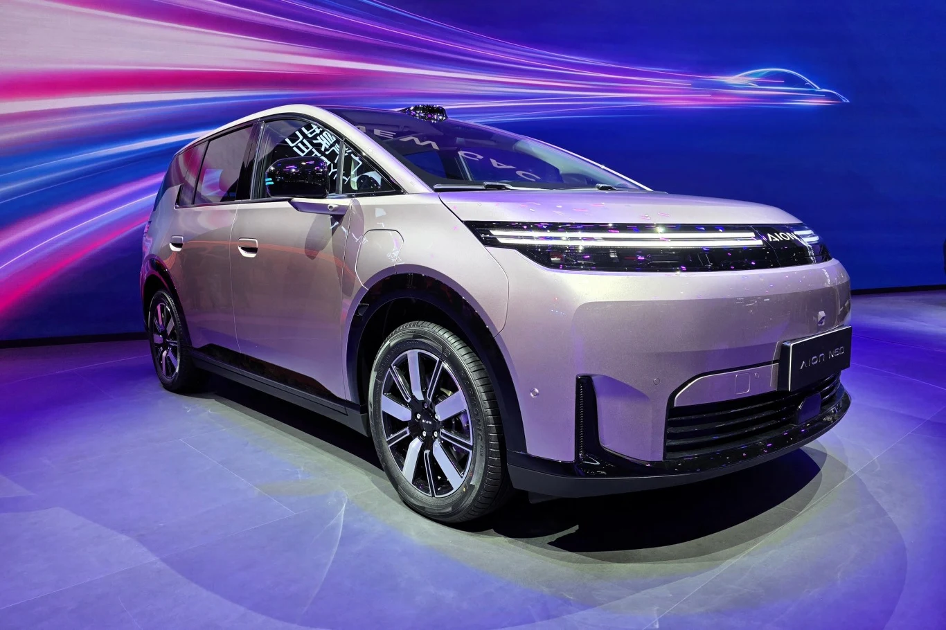 Nowoczesny minivan w metalicznym odcieniu różu z futurystyczną stylistyką przedniego pasa, wąskimi reflektorami LED i dużymi felgami, ustawiony na oświetlonej ekspozycji z dynamicznym, kolorowym tłem.