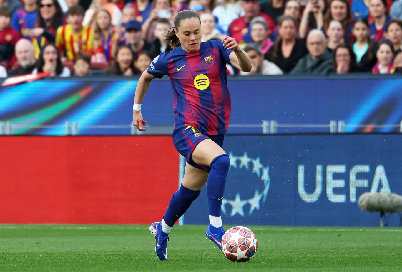 Piłkarka FC Barcelony w granatowo-bordowym stroju prowadzi piłkę na boisku podczas meczu, w tle widoczna publiczność i elementy oprawy UEFA.