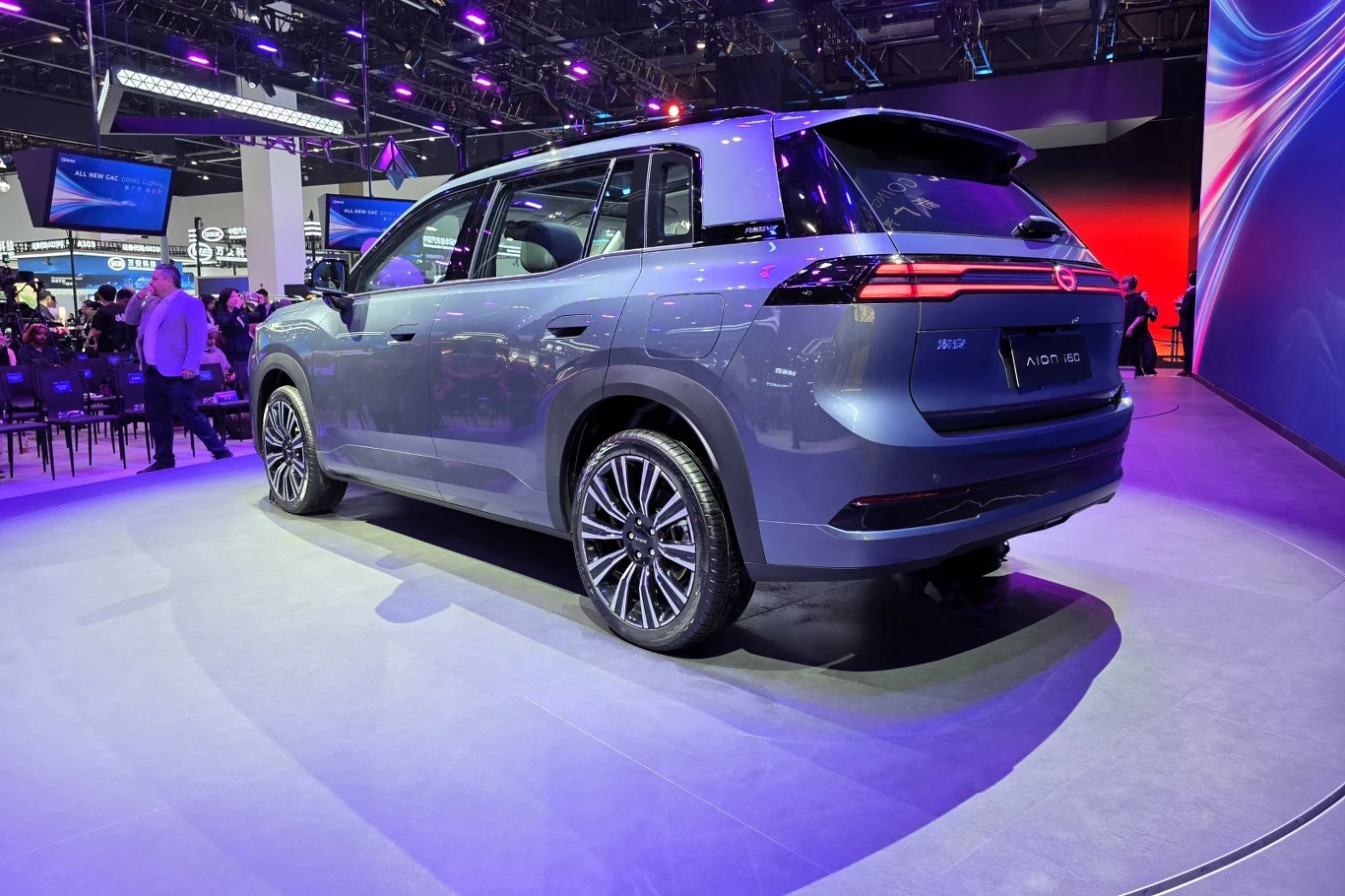 Nowoczesny, niebieskoszary SUV o dynamicznej sylwetce, podświetlony fioletowymi reflektorami podczas prezentacji na targach motoryzacyjnych, eksponuje charakterystyczne tylne światła LED oraz duże, aluminiowe felgi.