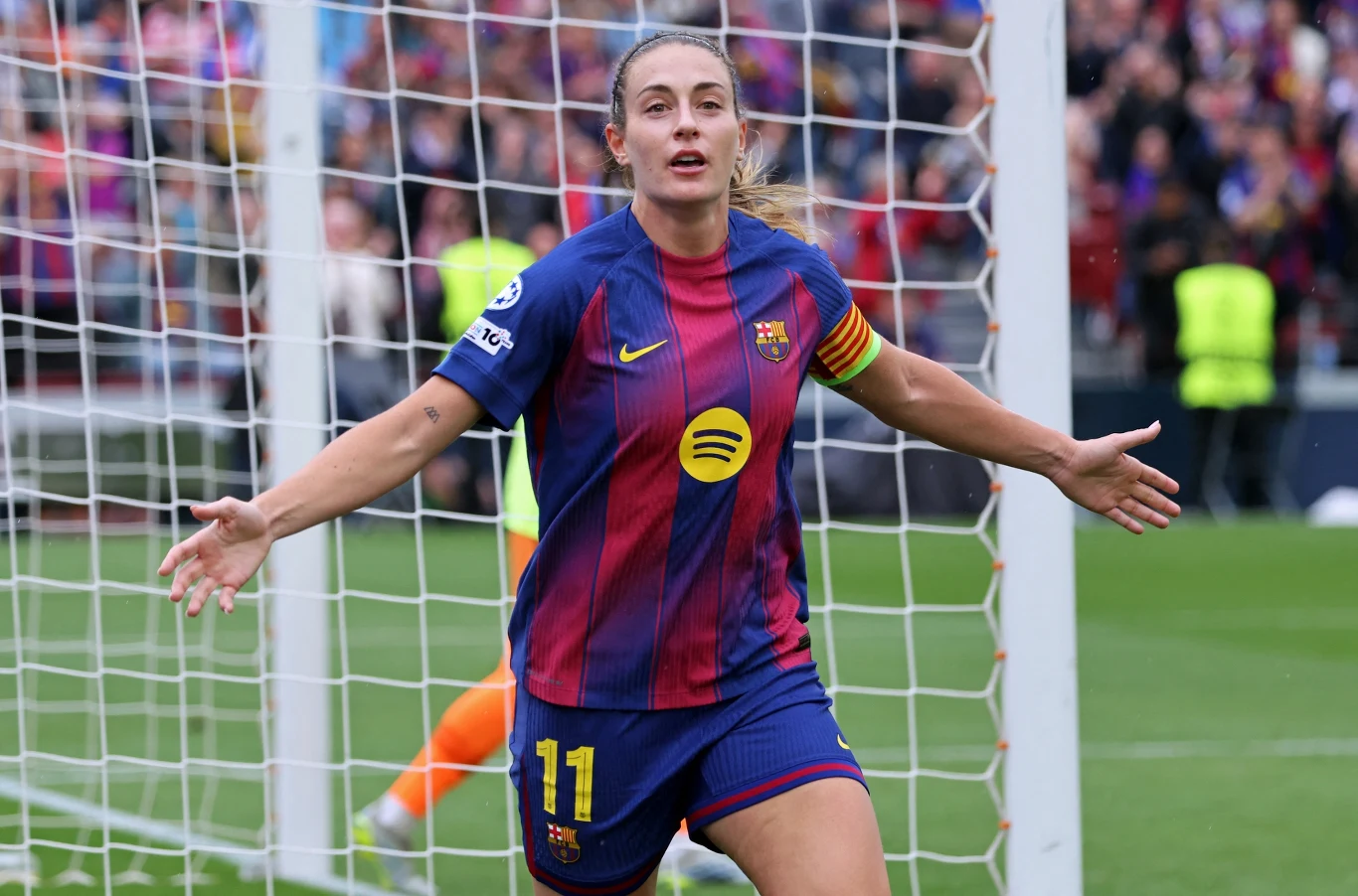 Piłkarka w stroju FC Barcelona z rozłożonymi ramionami świętuje zdobycie bramki na tle bramki piłkarskiej i siatki.