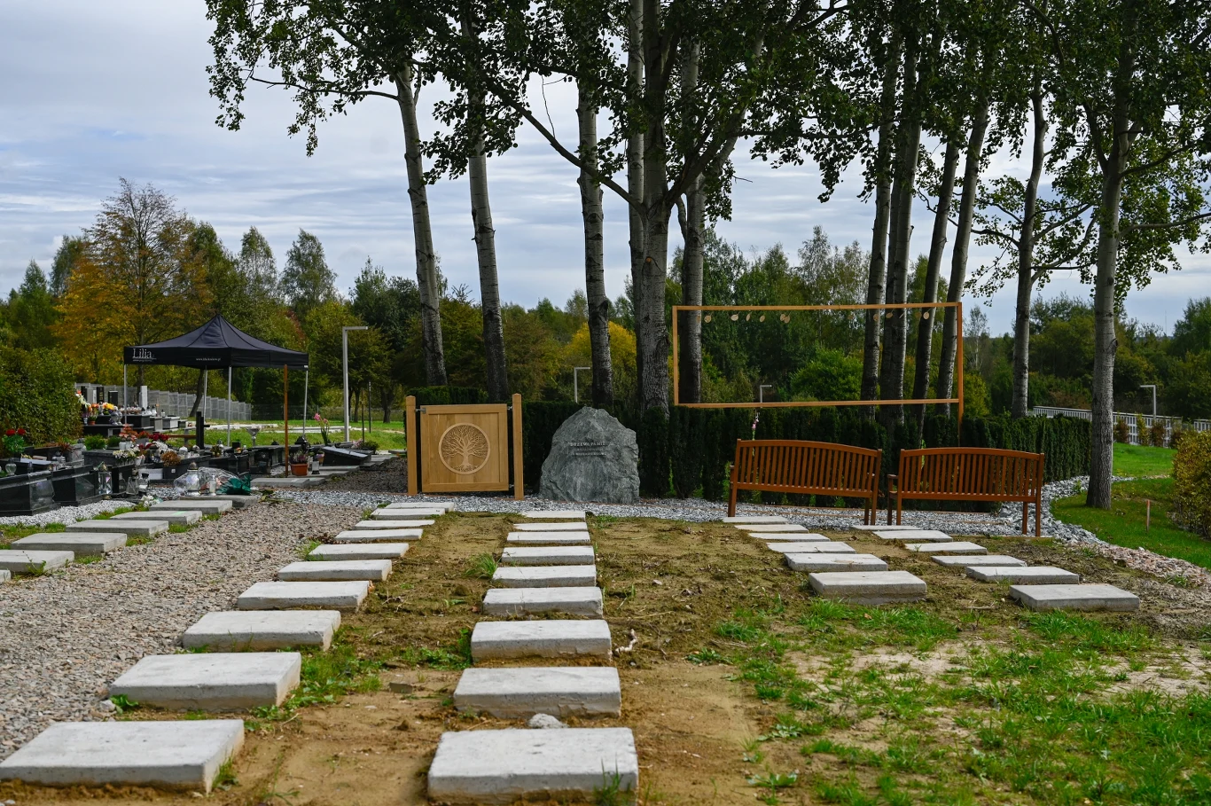 Cmentarz Podgórki Tynieckie Prostokątna alejka z betonowych płyt prowadzi do przestrzeni z kamieniem pamiątkowym otoczonym zielenią, po bokach ustawione są dwie drewniane ławki oraz drewniana konstrukcja ogrodowa.