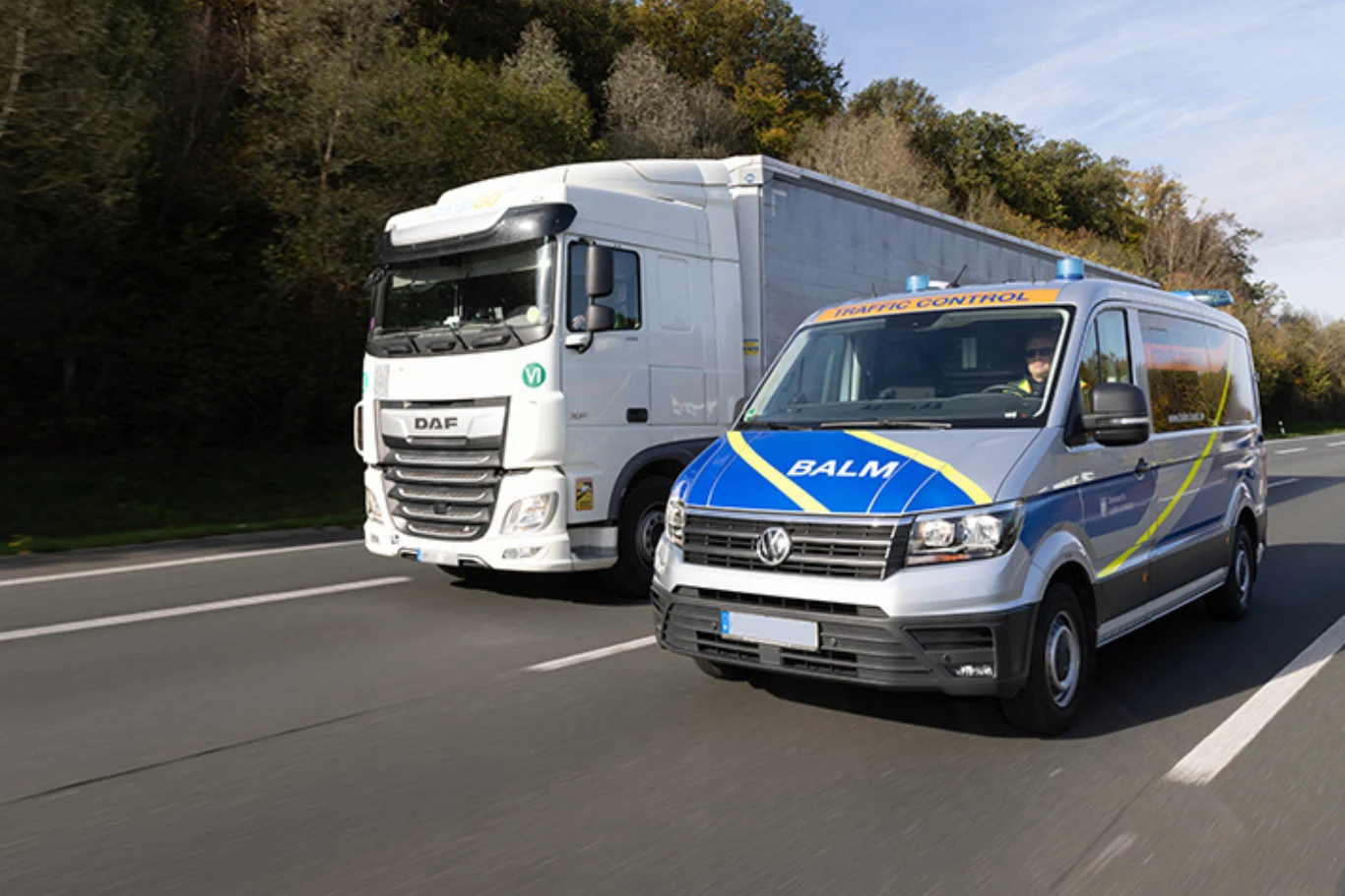 Jadący po autostradzie biały samochód ciężarowy marki DAF oraz oznakowany bus inspekcji transportu drogowego BALM, oba pojazdy poruszają się jednym kierunku, w otoczeniu drzew przy drodze.