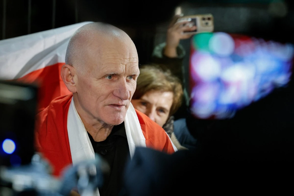Aleś Bialacki owinięty w biało-czerwoną flagę siedzi w otoczeniu innych osób, które robią mu zdjęcia i nagrywają telefonem.