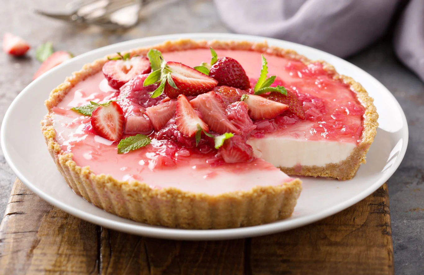 Ciasto z rabarbarem i truskawkami, galaretką i masą jogurtową Tarta z rabarbarem, truskawkami, masą jogurtową i galaretką, udekorowana truskawkami i miętą na białym talerzu.