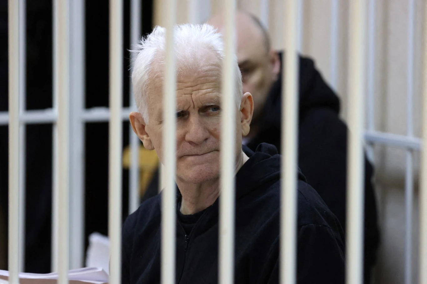 Aleś Bialacki siedzi w czarnej kurtce za metalowymi kratami, w tle widoczna inna osoba.