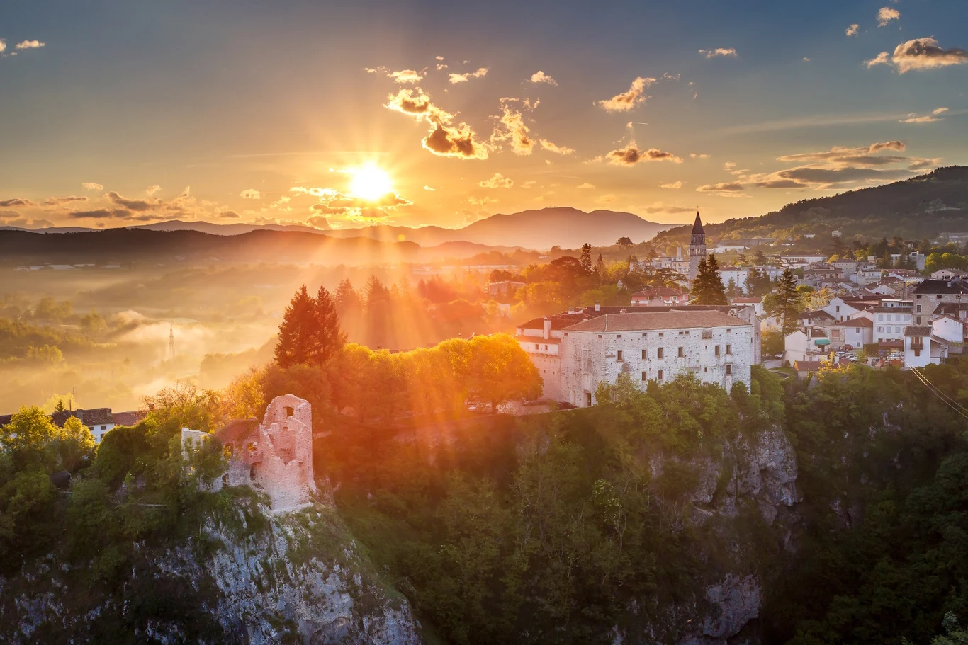 Ruiny średniowiecznego zamku położone na skalistym wzgórzu, otoczone gęstą zielenią, z panoramą miasta i gór w tle podczas zachodu słońca.