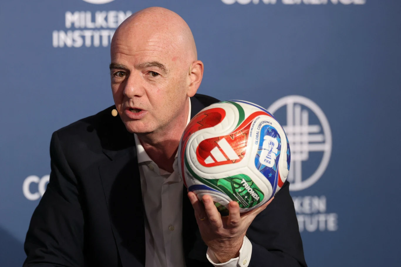 Gianni Infantino Mężczyzna w garniturze trzyma kolorową piłkę do piłki nożnej, mówi do mikrofonu przy uchu, w tle niebieska ścianka z logo Milken Institute.