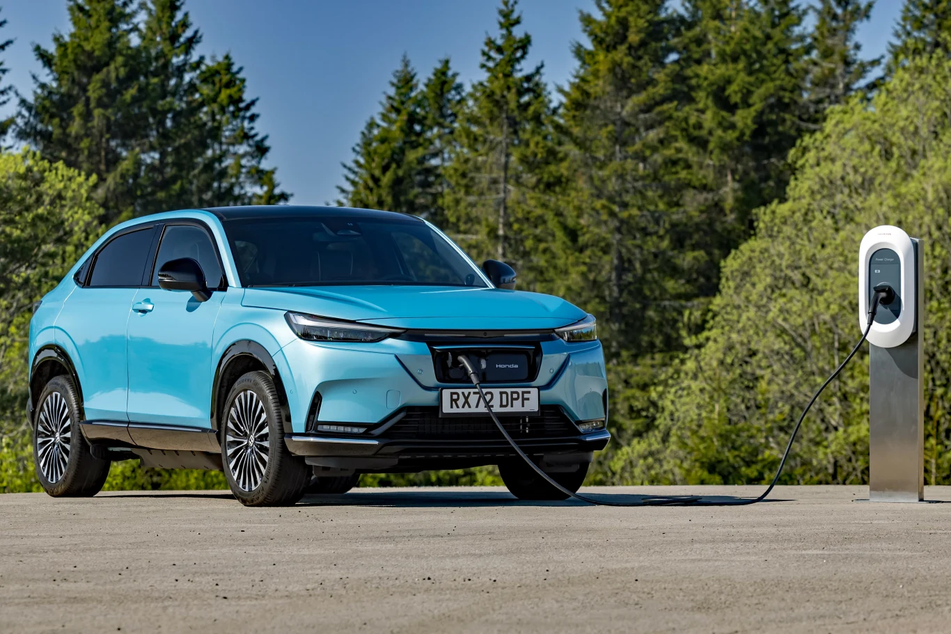 Niebieski samochód elektryczny marki Honda podłączony do stacji ładowania na tle zielonych drzew.