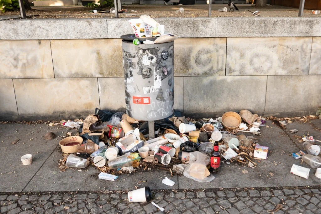 Przepełniony kosz na śmieci otoczony rozsypanymi wokół niego odpadkami, głównie papierowymi kubkami, butelkami i opakowaniami po jedzeniu.