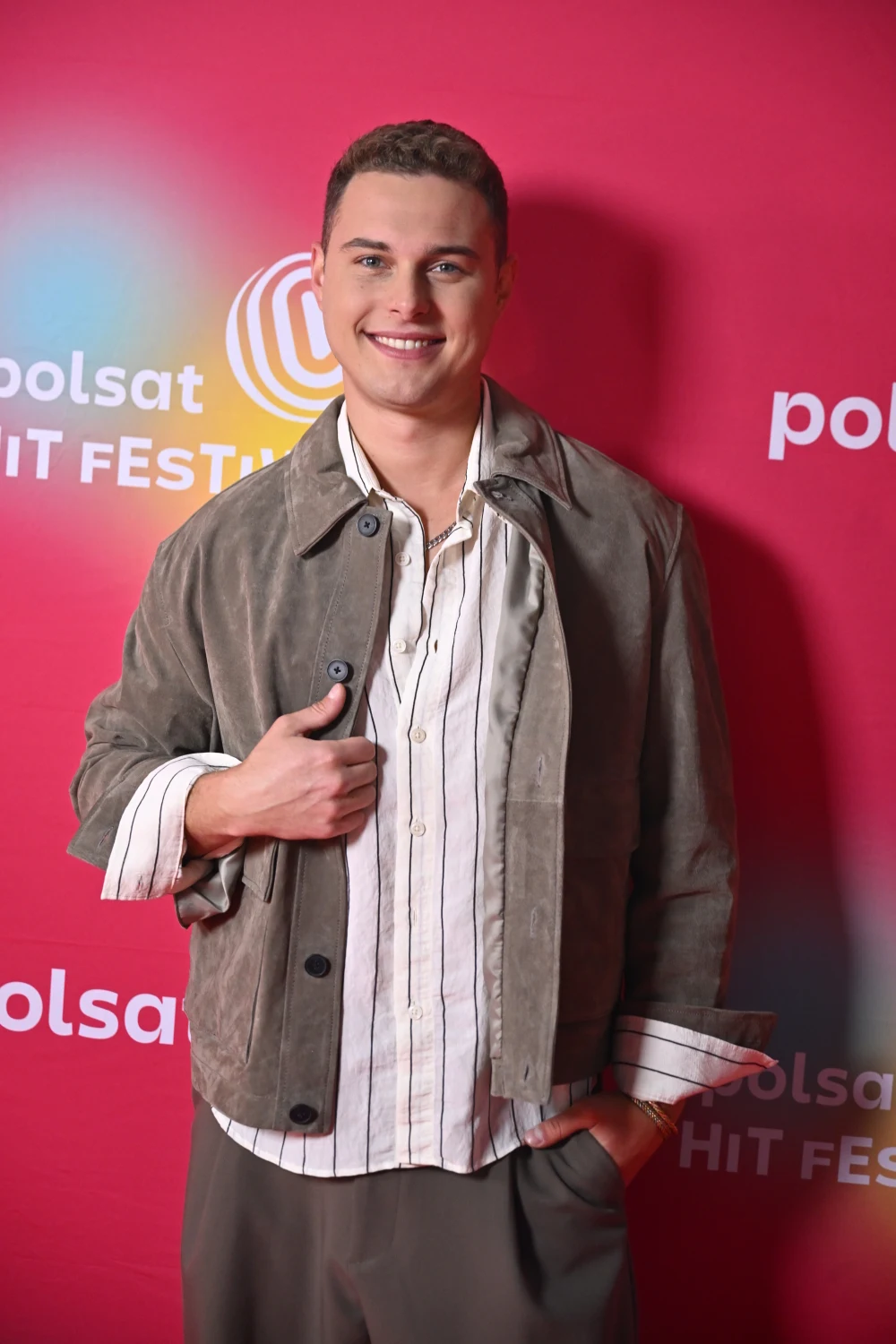 Uśmiechnięty mężczyzna w jasnej koszuli w paski i szarej kurtce pozuje na tle różowej ścianki z logo Polsat Hit Festiwal.