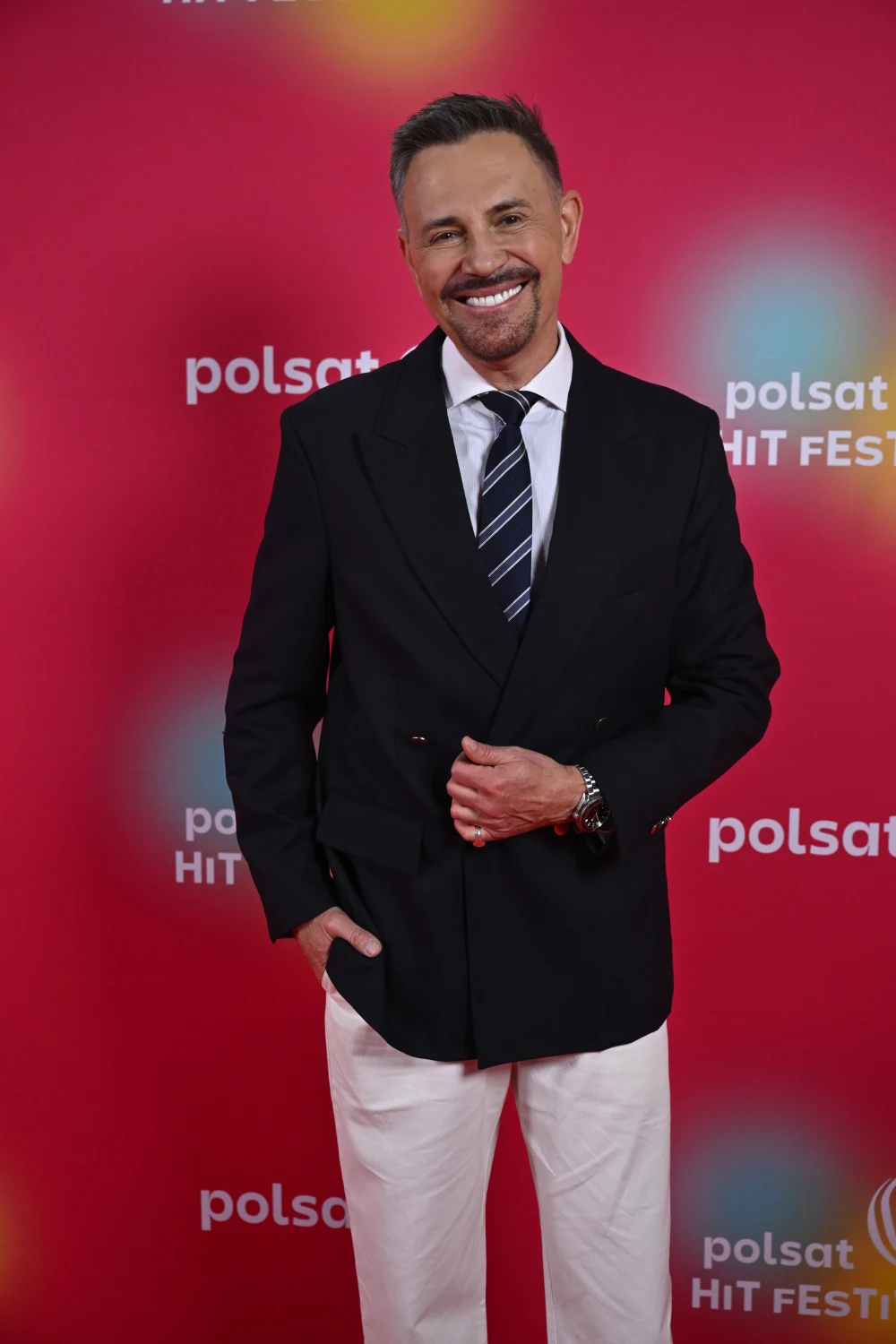 Mężczyzna w ciemnej marynarce, białej koszuli i krawacie w paski pozuje na tle ścianki z logo Polsat Hit Festiwal, uśmiechnięty i elegancki.