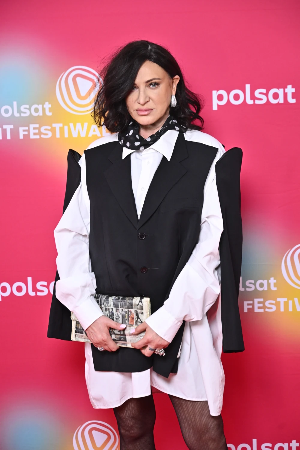 Kobieta z ciemnymi włosami ubrana w białą koszulę, oversize'ową czarną kamizelkę oraz czarną apaszkę w białe grochy, trzymająca kopertówkę na tle różowej ścianki z logotypami Polsat i Festiwal.