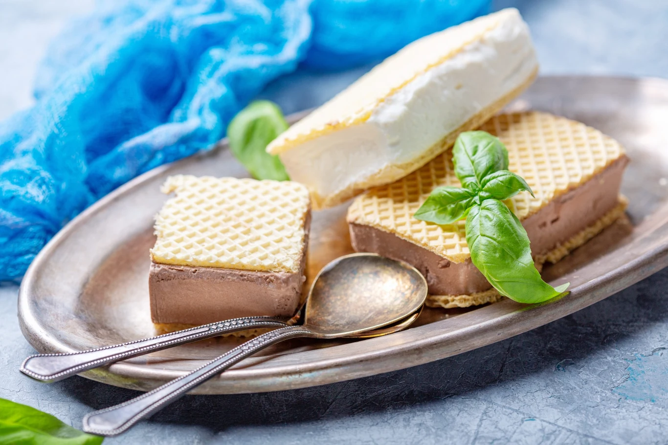 Lody w wafelku o dwóch smakach, podane na metalowej tacy z dwiema łyżeczkami i świeżymi listkami bazylii.