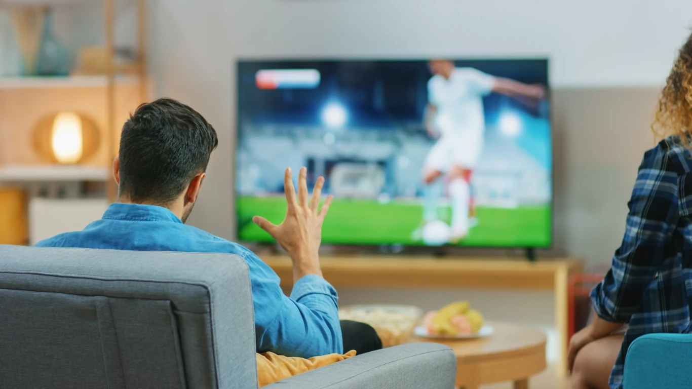 Telewizja internetowa się zacina. Jak uratować mecz albo serial? Mężczyzna gestykuluje do zacinającego się telewizora z meczem piłki nożnej, obok siedzi kobieta na kanapie.