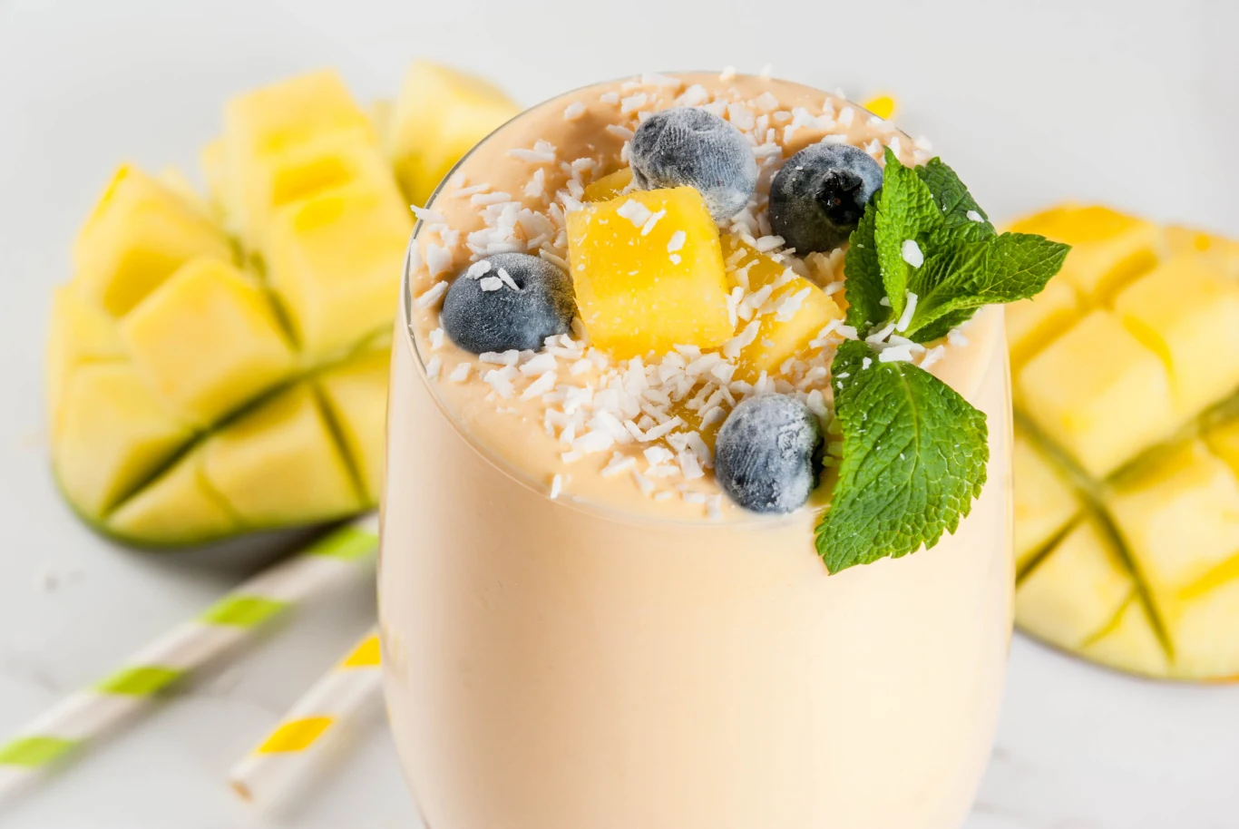 Mango uznawane jest za jeden z najbardziej wartościowych owoców. Szklanka kremowego smoothie z mango, udekorowana kostkami mango, jagodami, wiórkami kokosowymi i świeżą miętą, w tle połówki pokrojonego mango.