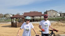 Dwie osoby w kaskach rowerowych i sportowych okularach stojące obok swoich rowerów górskich na brukowanym placu, w tle drewniana altana, piasek oraz budynki mieszkalne.