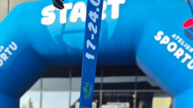 Medal zawieszony na niebieskiej tasiemce z napisem, na tle dużej niebieskiej bramy z napisem 'START' i stojącego przed wejściem roweru.
