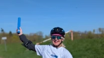 Kobieta w sportowym stroju jadąca rowerem po utwardzonej ścieżce, uśmiechnięta, z niebieską pałeczką w uniesionej ręce i kaskiem na głowie, zielony wał i niebieskie niebo w tle.