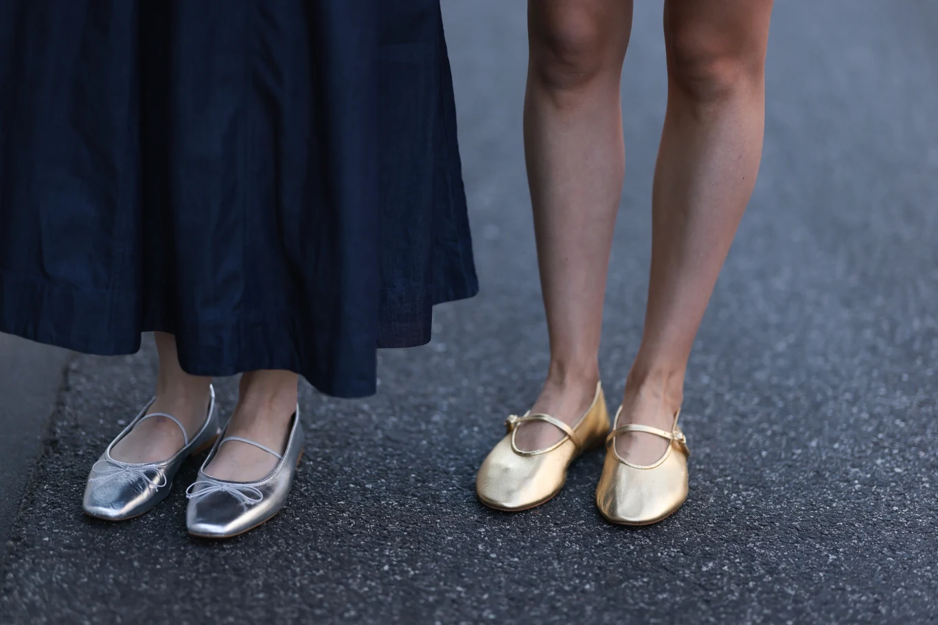 Modne buty na lato 2026 Dwie osoby stojące na asfalcie, jedna w srebrnych balerinach i długiej, ciemnej spódnicy, druga w złotych balerinach z odkrytymi nogami.