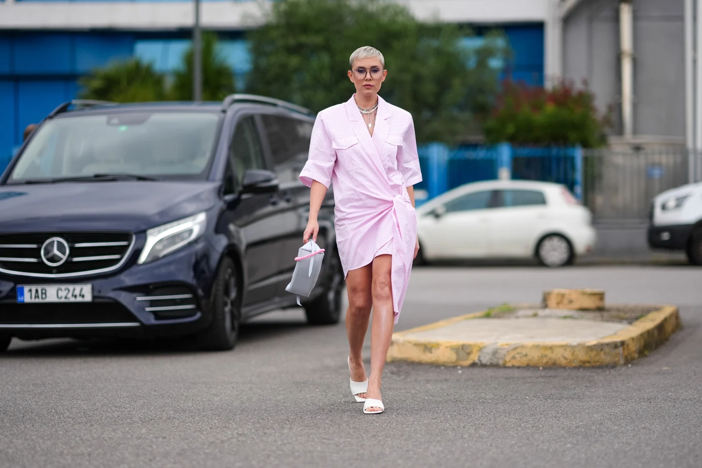 Kobieta z krótkimi blond włosami ubrana w różową sukienkę koszulową i białe sandały, trzymająca szarą torebkę, idzie po miejskim parkingu obok czarnego samochodu osobowego marki Mercedes.