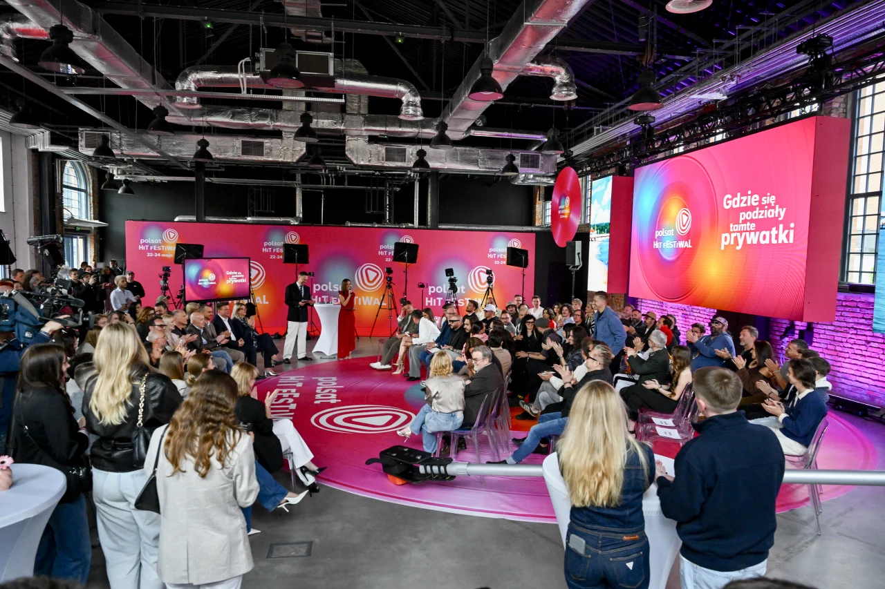 Grupa osób zgromadzona w nowoczesnej sali eventowej z dużymi ekranami i sceną, na których dominuje kolor różowy i motyw medialny; uczestnicy oglądają prezentację dotyczącą programu rozrywkowego, część z nich siedzi, inni stoją, wokół rozstawiony profes...