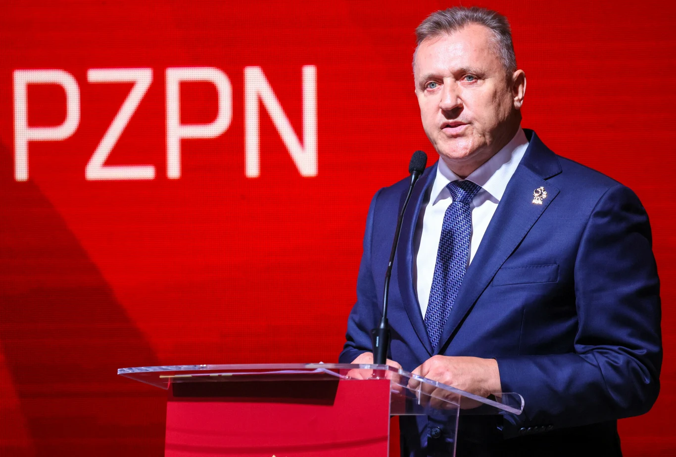 Mężczyzna w eleganckim garniturze przemawia przy mównicy na tle czerwonej ściany z dużym napisem PZPN.