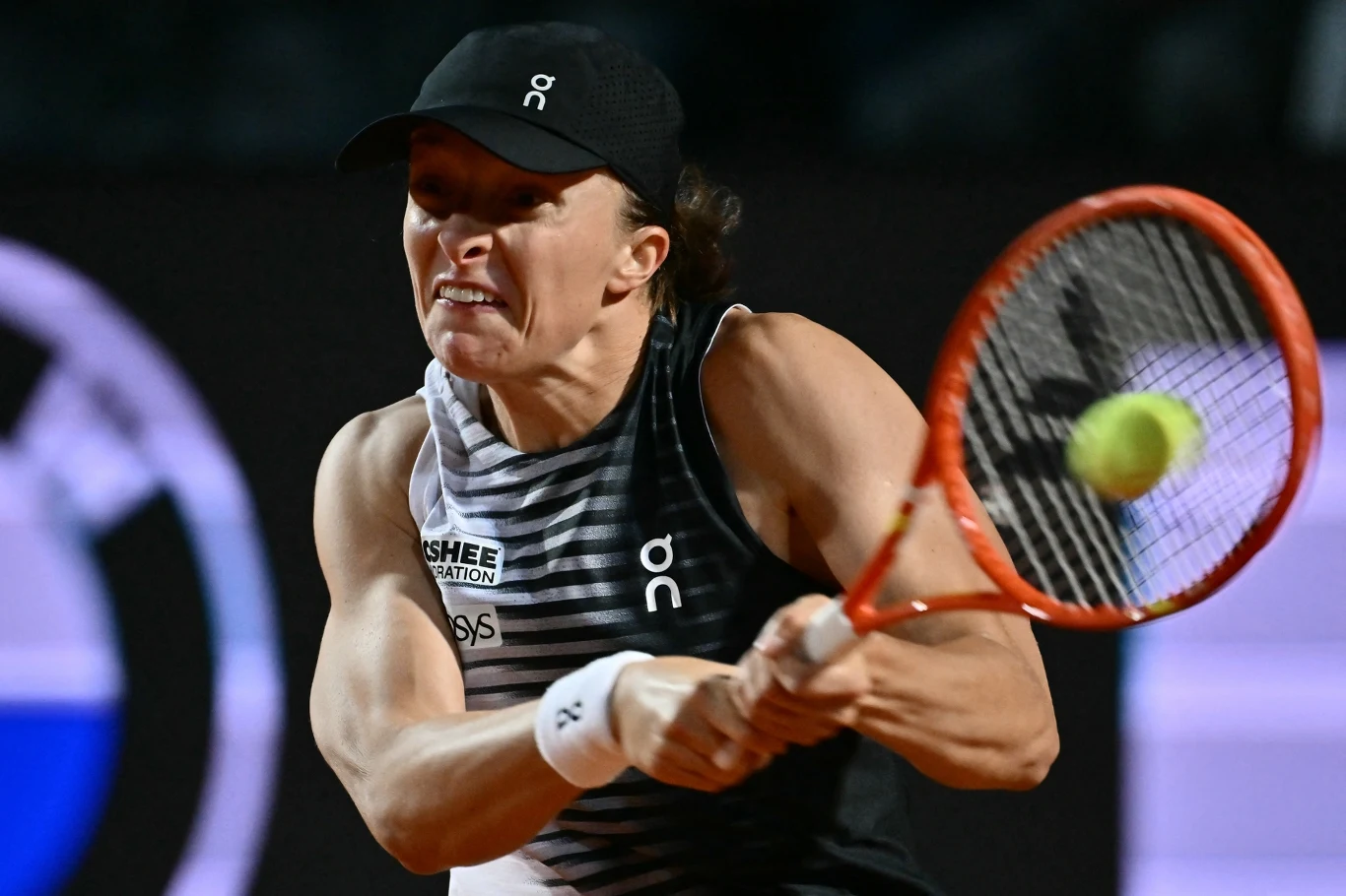 Tenisistka w ciemnej czapce i stroju sportowym wykonuje dynamiczny backhand, skupiona na uderzeniu piłki tenisowej rakietą.