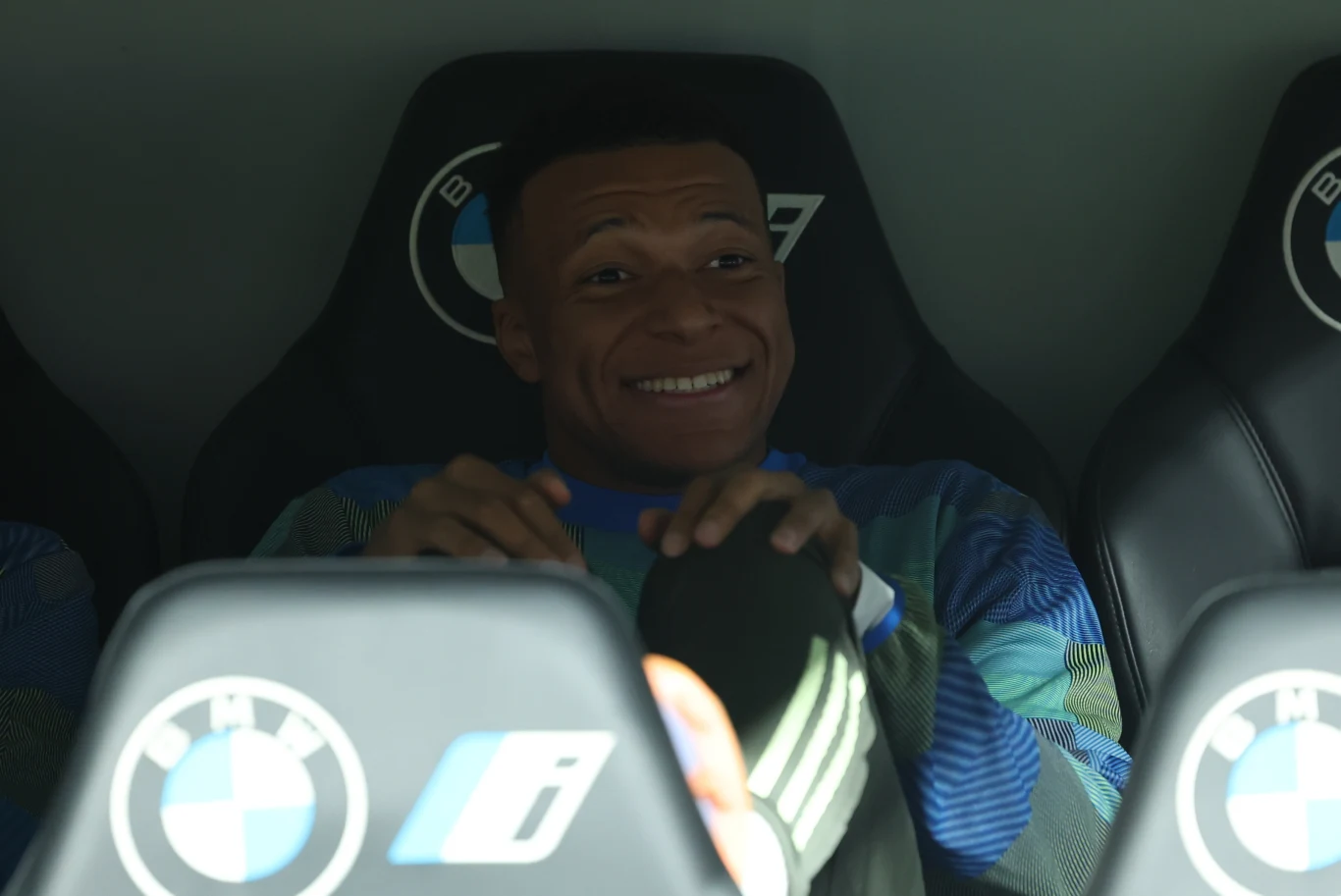 Uśmiechnięty młody mężczyzna w sportowej bluzie siedzi na fotelu z logo BMW, opierając dłonie na nogach.