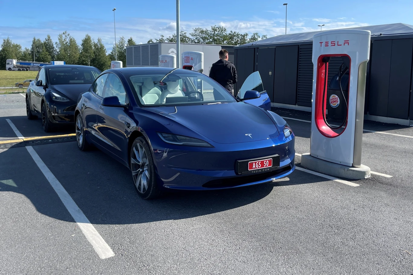 Tesla testuje na wybranych Superchargerach nową funkcję. Lista kolejkowa ogarnie chaos przy ładowarkach Niebieska Tesla ładowana na stacji Supercharger, z tyłu czarny samochód i osoba przy ładowarce.