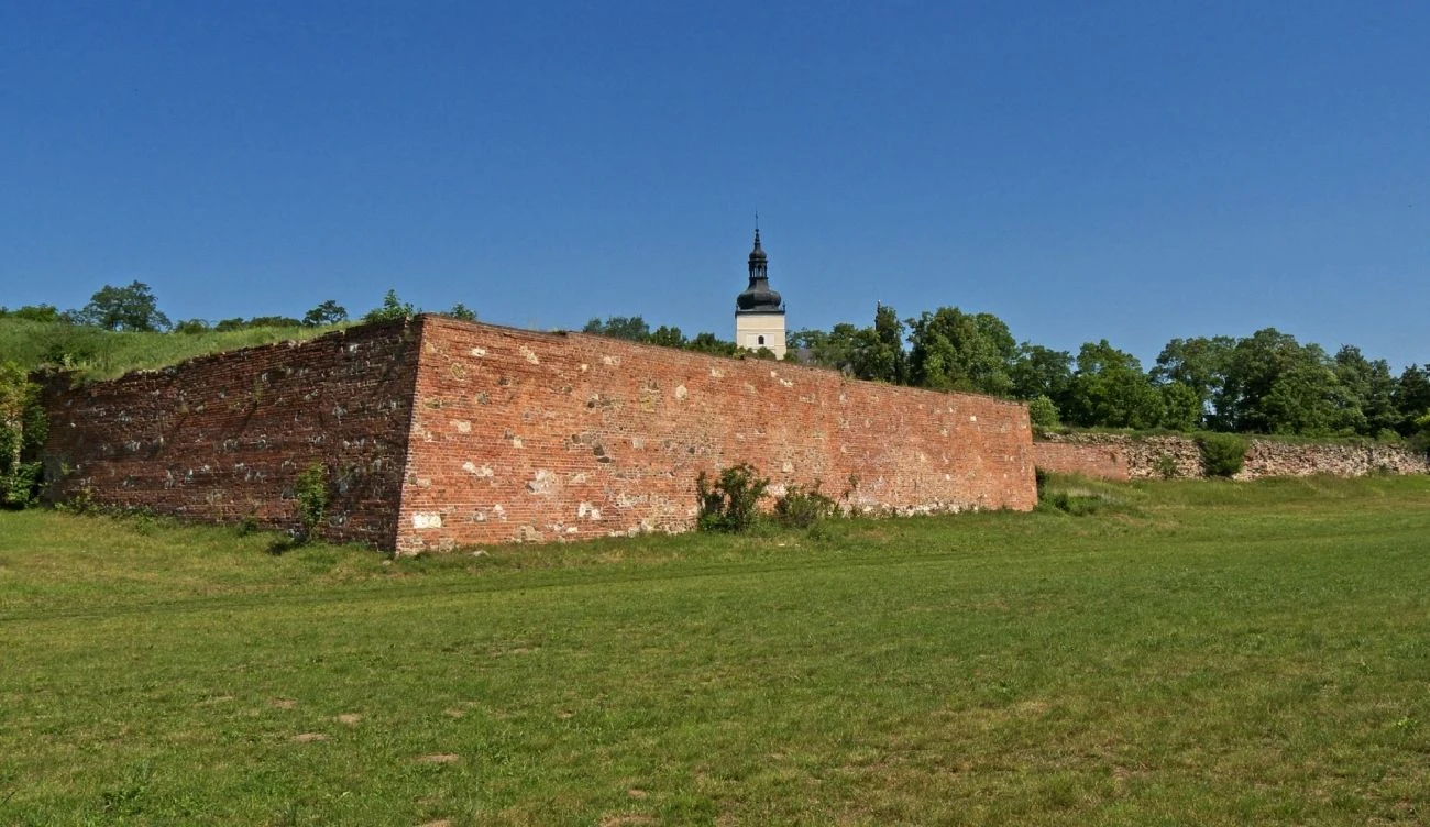 Ceglany fragment muru na tle zielonej trawy, w oddali wieża kościelna otoczona drzewami pod błękitnym niebem.