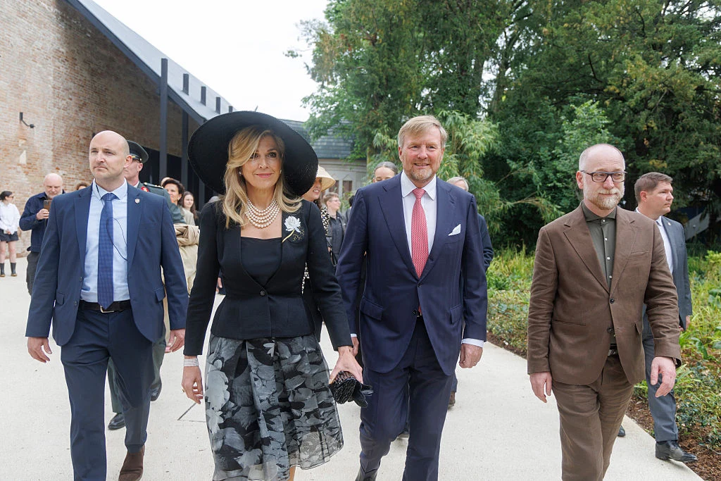 Cztery elegancko ubrane osoby idące razem, na czele kobieta w czarnym kapeluszu i mężczyzna w granatowym garniturze z różowym krawatem, w tle zieleń i fragment budynku o nowoczesnej architekturze.