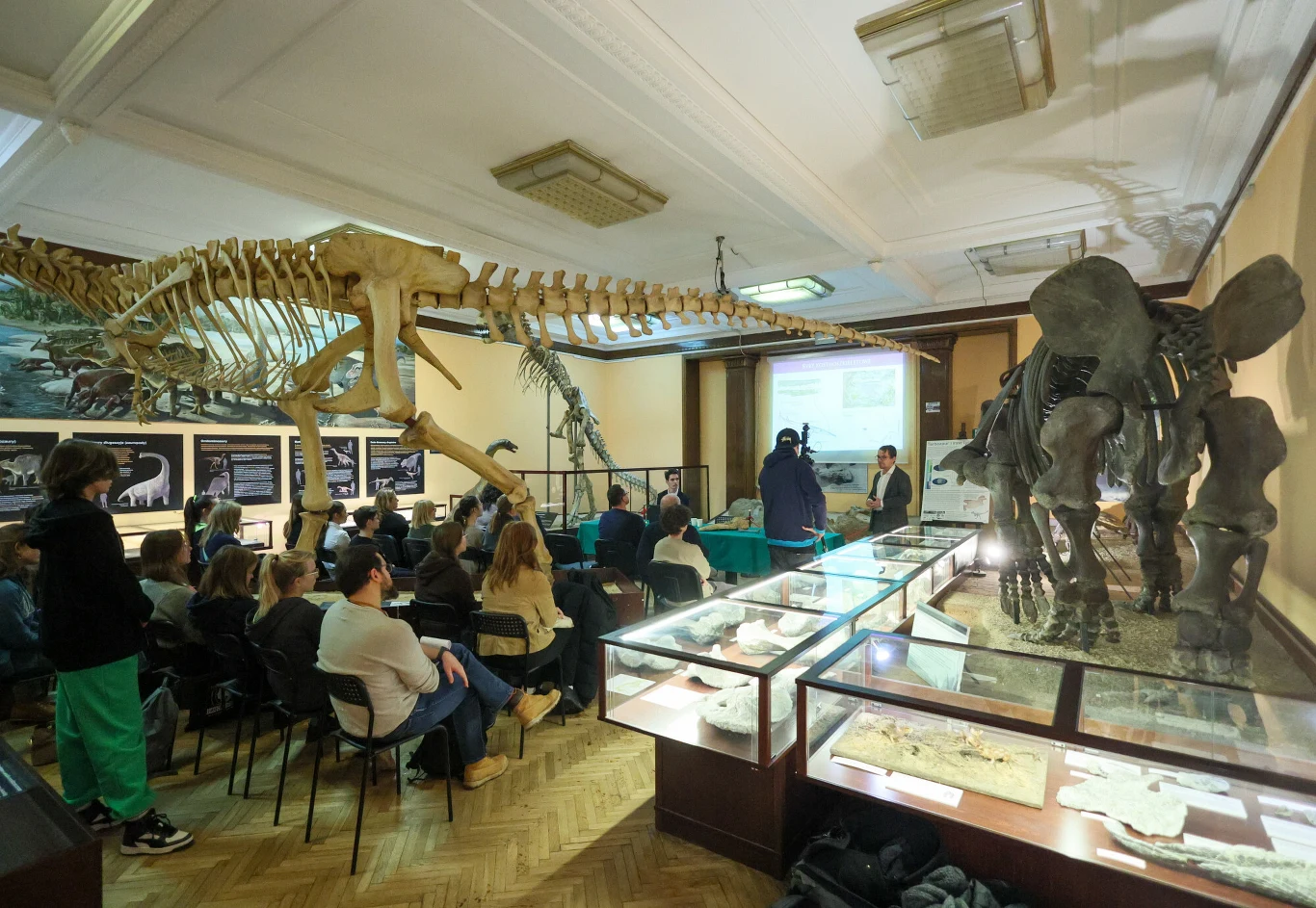 Sala muzealna z dużymi szkieletami dinozaurów, gablotami z eksponatami oraz grupą osób słuchających prelekcji.