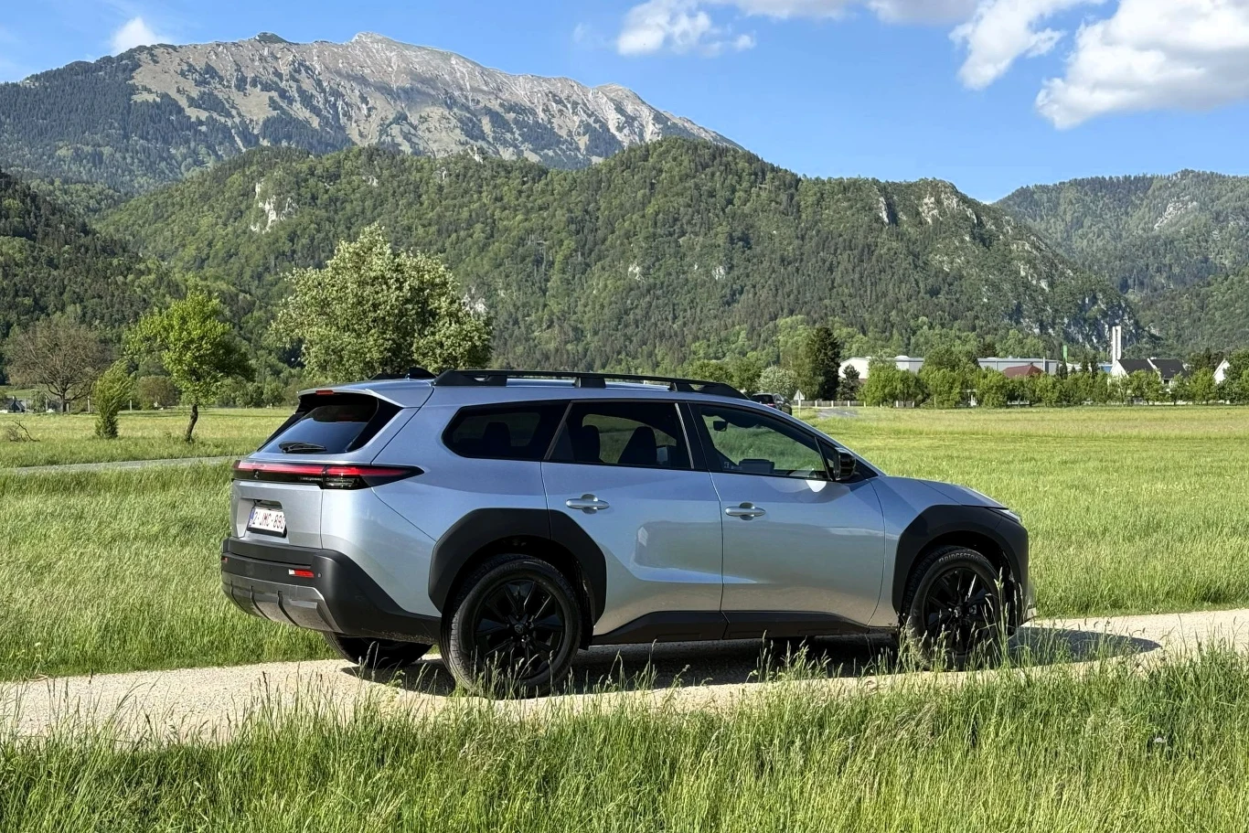 Srebrny SUV o nowoczesnym designie stoi na wiejskiej drodze, otoczony zielonymi polami i górami w tle.
