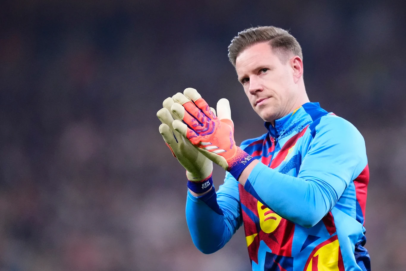 Marc-Andre ter Stegen Bramkarz w kolorowej bluzie sportowej i rękawicach, stojący na boisku i składający dłonie w geście podziękowania.