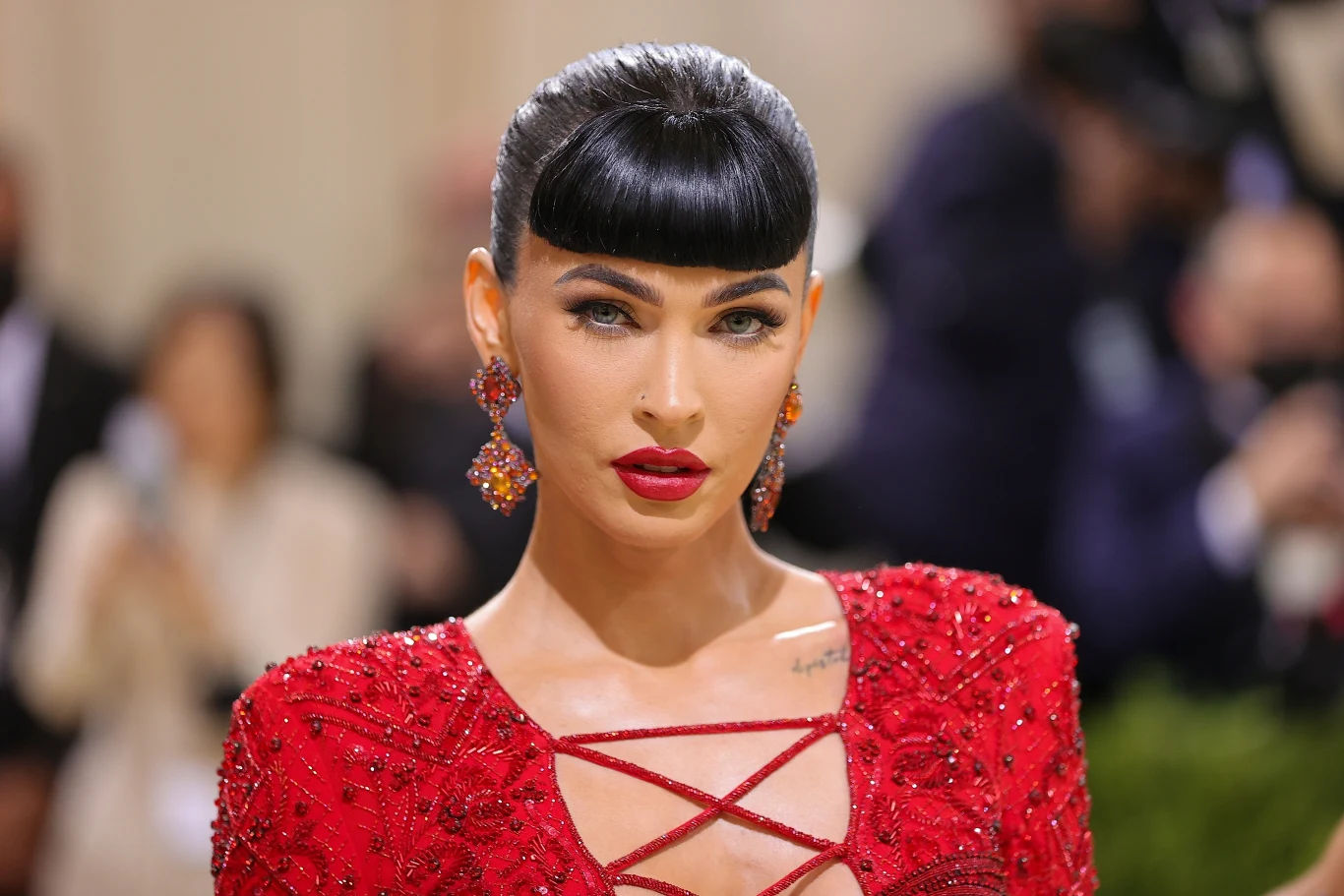 Megan Fox w 2021 roku Kobieta z ciemnymi, spiętymi włosami i prostą grzywką, ubrana w efektowną czerwoną suknię z koralikami oraz czerwone kolczyki, na tle rozmytego, eleganckiego wnętrza.