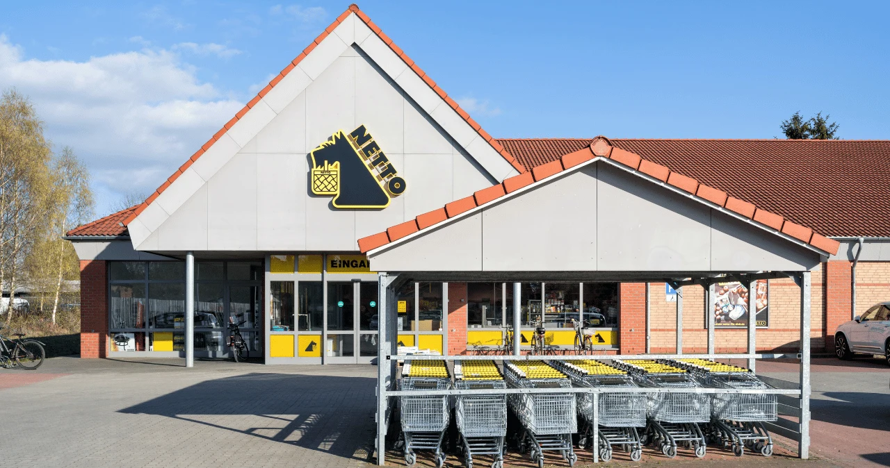 Netto kończy akcję 1+1 gratis. Jeszcze dziś kupisz kawę i Persil taniej Wejście do supermarketu Netto z logo i rzędem wózków zakupowych przed budynkiem