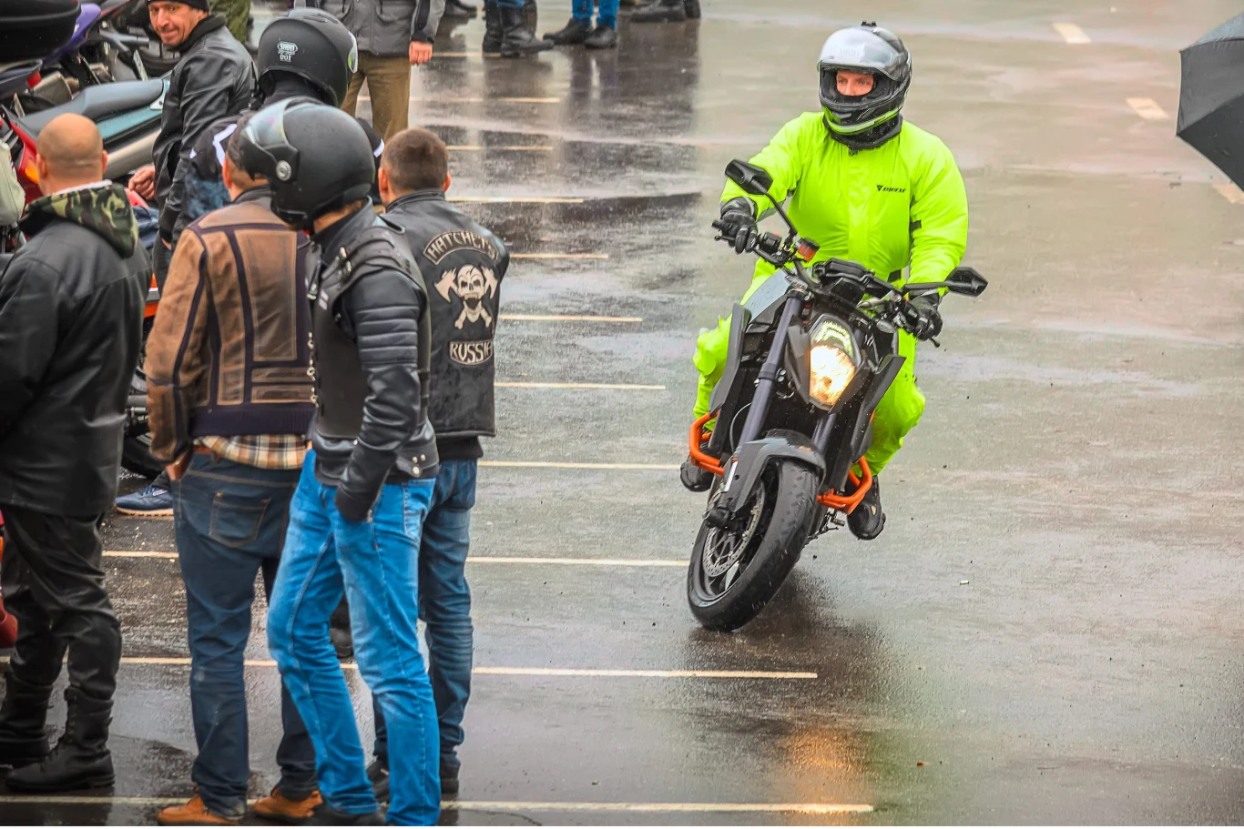 Motocyklista w jaskrawozielonym kombinezonie jedzie po mokrym asfalcie na czarnym motocyklu, mijając grupę osób ubranych w kurtki skórzane i kaski motocyklowe.