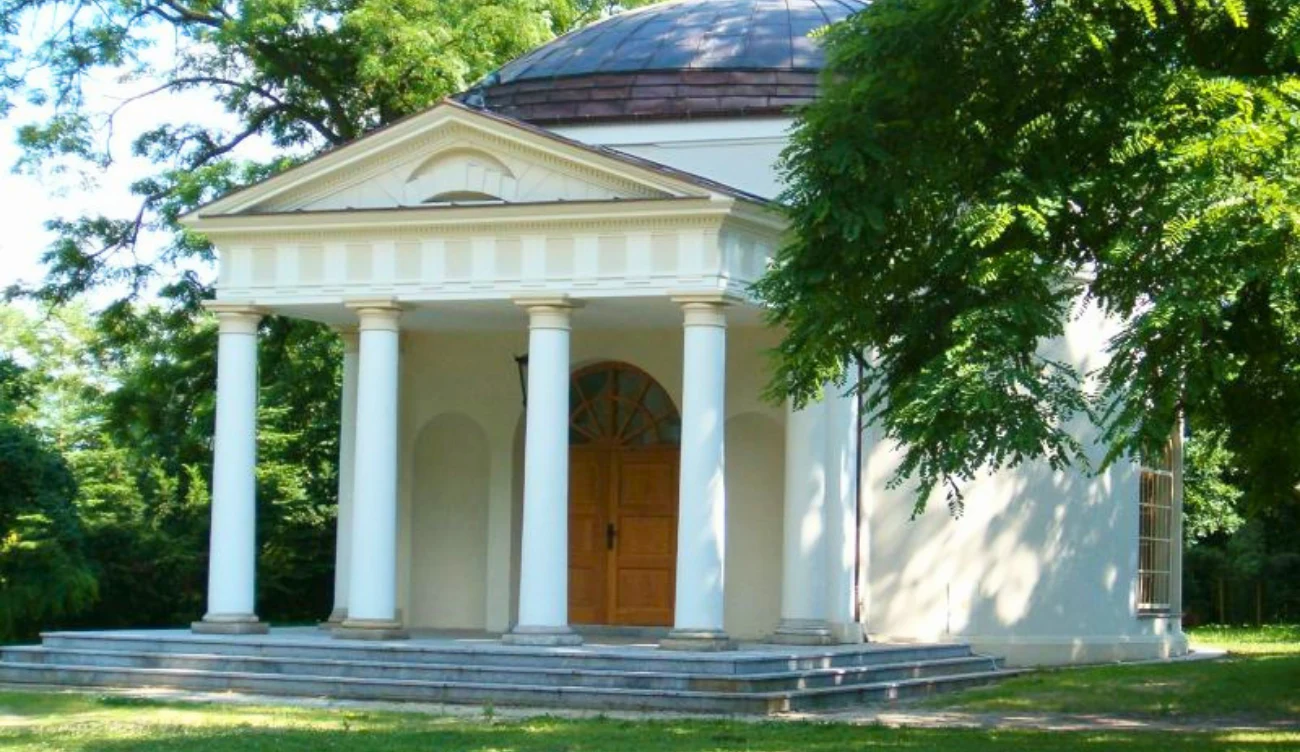 Klasycystyczny budynek z kolumnowym portykiem, centralnie usytuowanymi drewnianymi drzwiami oraz kopułą otoczony zielenią drzew.