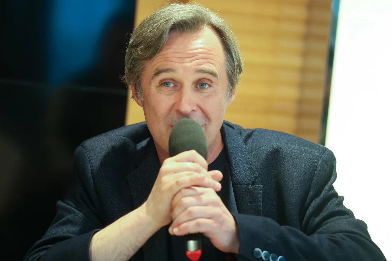 Mężczyzna w średnim wieku w granatowej marynarce trzyma mikrofon i mówi, patrząc w kierunku obiektywu.
