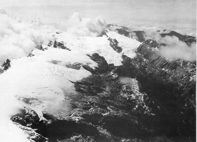Pokrywa lodowa Puncak Jaya 1936 Ośnieżone szczyty gór rozciągające się wśród chmur, strome zbocza i lodowce kontrastujące z ciemnymi skałami.