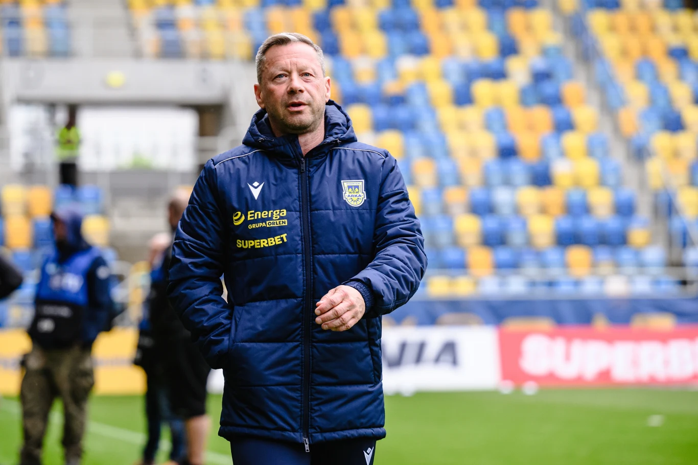 Trener drużyny piłkarskiej ubrany w granatową kurtkę z herbem klubu Arka Gdynia przechadza się po stadionie z żółto-niebieskim układem krzeseł na trybunach.