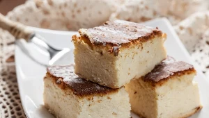 Trzy kawałki puszystego sernika z delikatnie przypieczoną, złotą skórką, ułożone na białym talerzu obok srebrnego widelca.