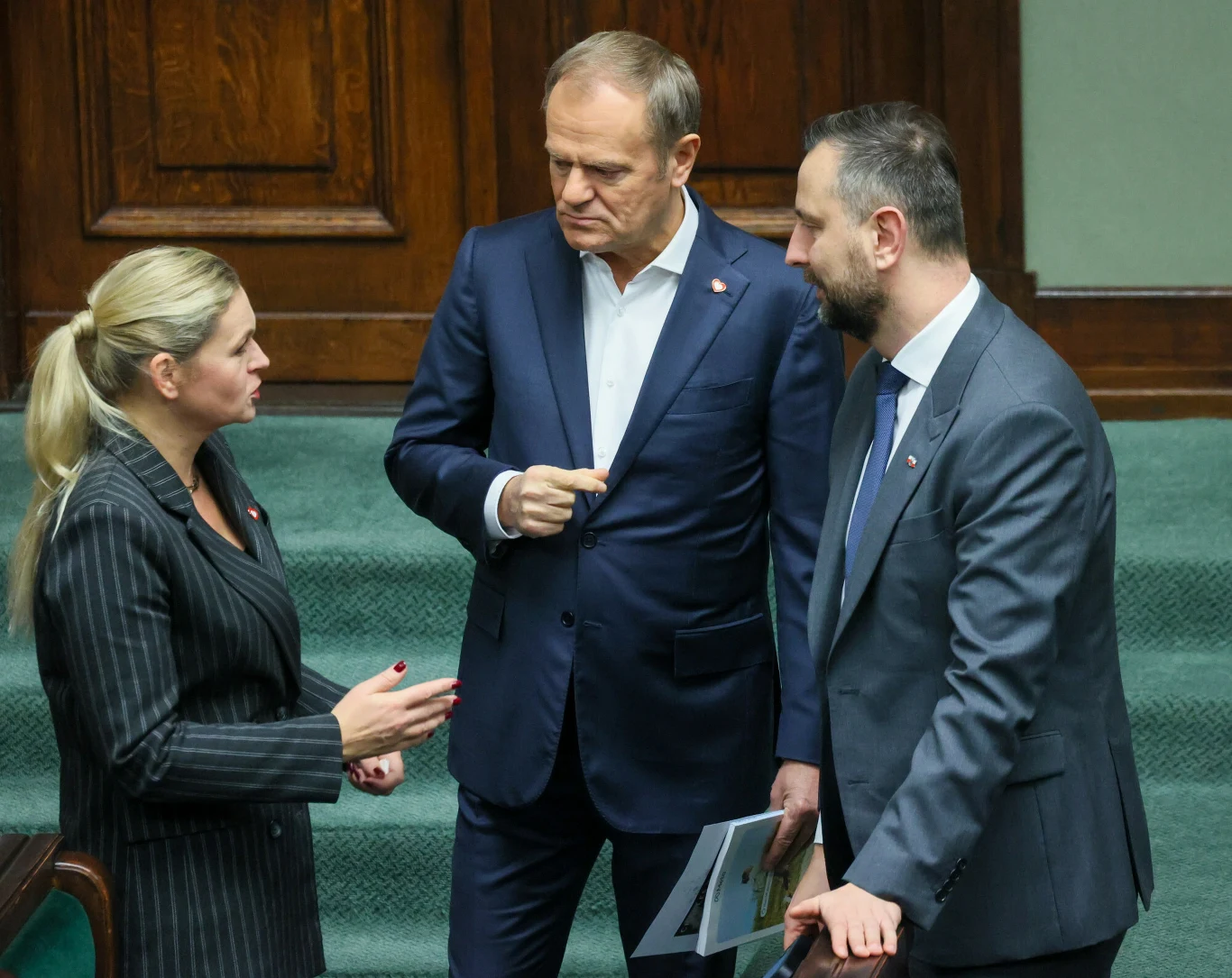 Trójka polityków w garniturach prowadzi poważną rozmowę na tle sejmowych ławek.