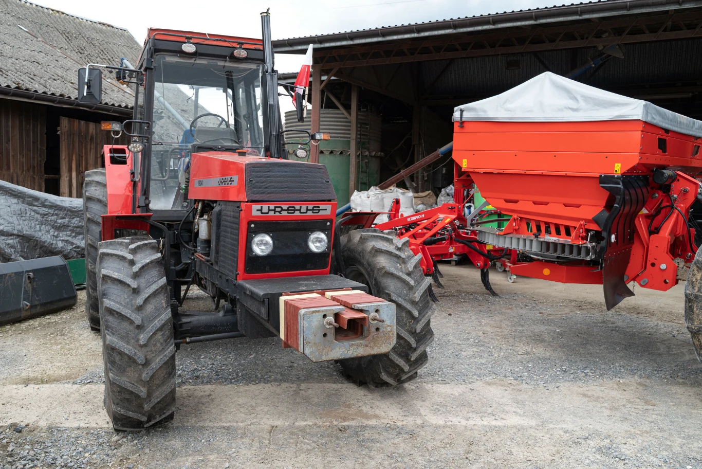 Czerwony traktor Ursus z dużym siewnikiem zaczepionym z tyłu stoi na podwórzu gospodarstwa, otoczony zabudowaniami gospodarczymi.